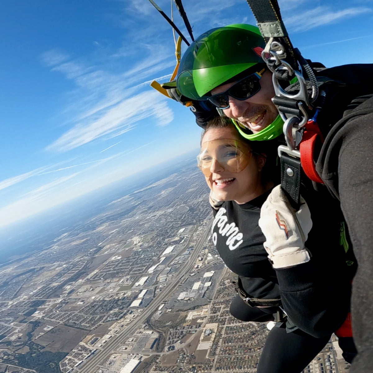 Two people smiling mid-air while steering a parachute during a tandem skydive